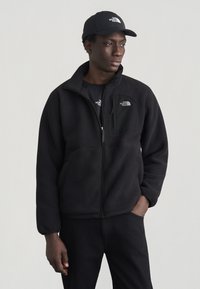 Άνδρας που φορά μαύρο φλις μπουφάν, μαύρο καπέλο και μαύρο παντελόνι, στέκεται με το ένα χέρι στην τσέπη, κοιτάζοντας προς το πλάι μπροστά σε ένα απλό φόντο.