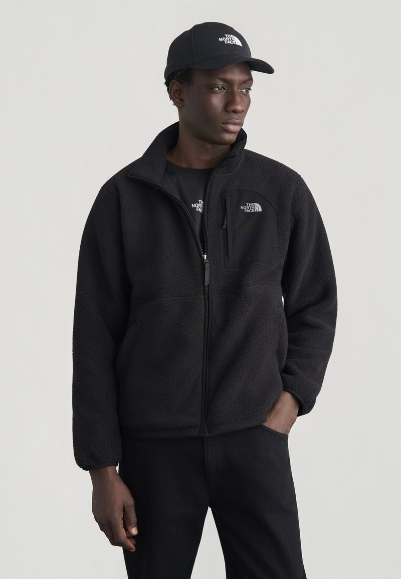 Άνδρας που φορά μαύρο φλις μπουφάν, μαύρο καπέλο και μαύρο παντελόνι, στέκεται με το ένα χέρι στην τσέπη, κοιτάζοντας προς το πλάι μπροστά σε ένα απλό φόντο.