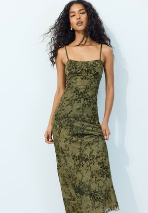 Femme aux longs cheveux noirs bouclés portant une robe midi ajustée fleurie vert olive avec de fines bretelles sur un fond clair uni.
