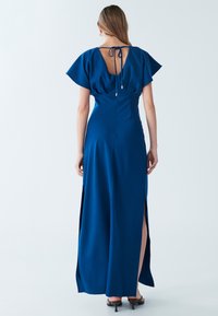 Dlhé, tmavomodré šaty s jemne plyskačkovými rukávmi, viazaným chrbtom a bočnými rozparkami. Vyrobené z hladkej látky, zvýrazňujúce priliehavý strih.