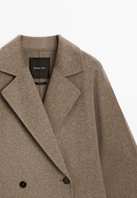 Bruine wollen jas met een dubbelgebreid ontwerp, scherpe revers en twee zwarte knopen. Etiket leest "Massimo Dutti." Zachte textuur.