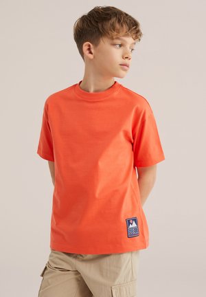 Rövid barna hajú fiú, aki narancssárga pólót visel kis logóval és bézs színű cargo rövidnadrágot, oldalra néz.
