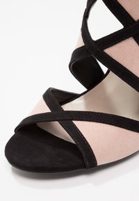 Gros plan d’une sandale à talon haut ouverte pour femme avec des brides en daim noir sur une base en tissu beige clair.