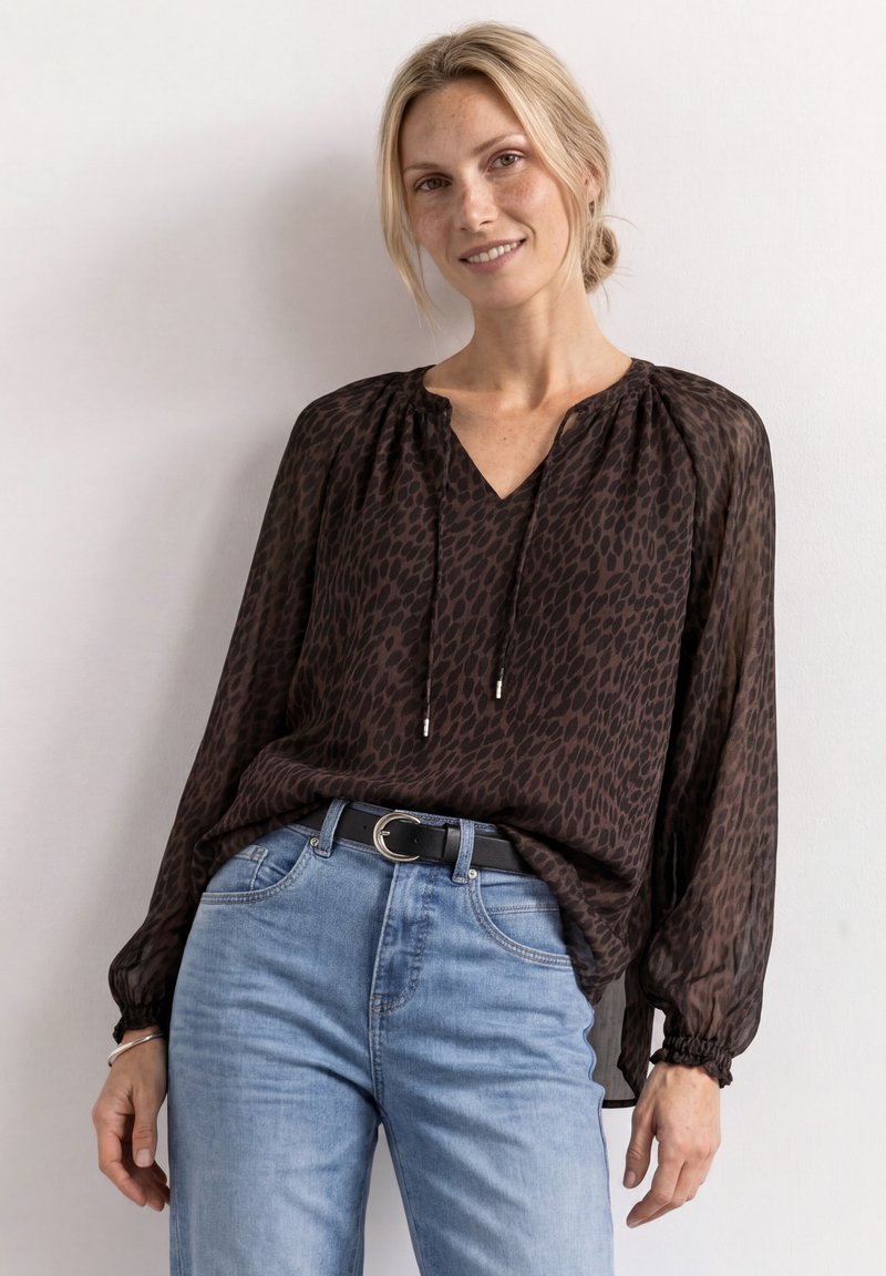 Femme blonde sourit, portant un chemisier transparent foncé à motifs avec un col à cordon et un jean bleu clair avec une ceinture noire, debout devant un mur blanc.