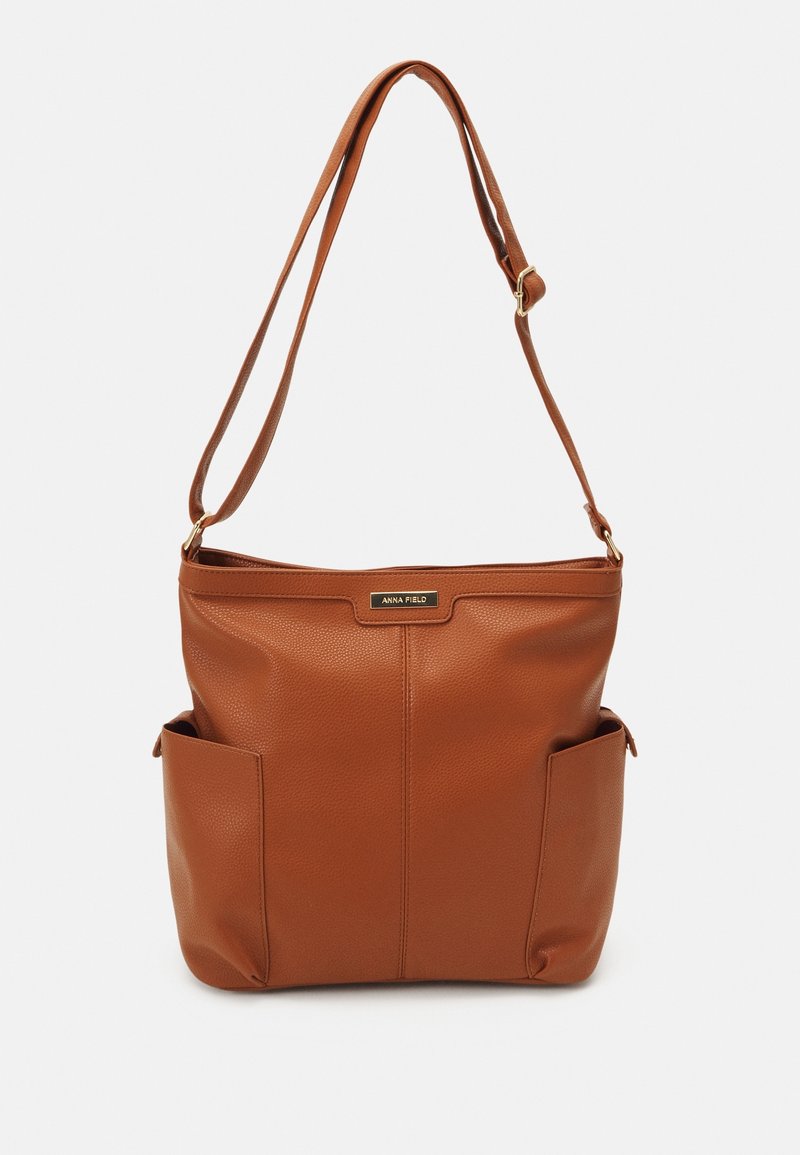 Sac à bandoulière en cuir marron avec sangle réglable, deux poches latérales et une petite étiquette métallique de marque au centre avant.