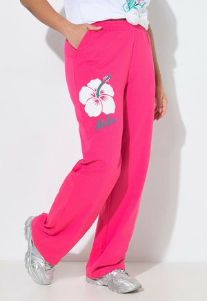 Person iført knaldpink bukser med en hvid hibiscusblomst og teksten "Aloha", kombineret med sølv sneakers og en hvid knudet T-shirt.