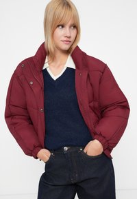 Chaqueta de plumas burdeos con cuello alto, cierre a presión, superficie texturizada, llevada sobre un suéter azul marino y pantalones de mezclilla oscuros.