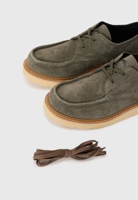 Olive green suede shoes with a round toe, brown rubber sole, and stitched detailing. Includes a matching brown shoelace beside them.