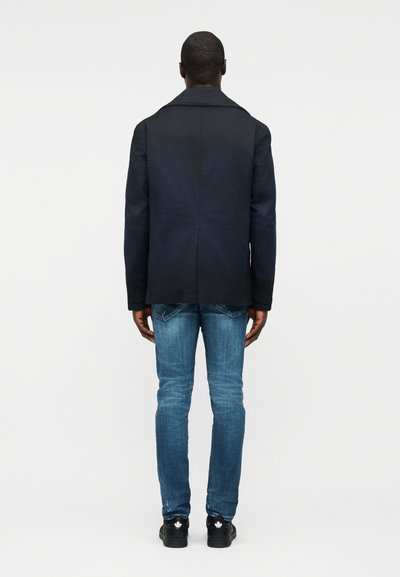 Homme vu de dos portant une veste noire, un jean bleu et des baskets noires avec des motifs de feuilles d'érable blanches.