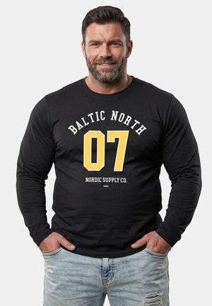 Lächelnder Mann mit dunklem Haar und Bart, der ein schwarzes Langarmshirt mit "Baltic North 07 Nordic Supply Co." und hellblauen zerrissenen Jeans trägt.
