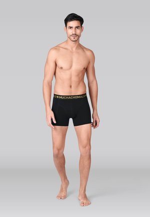 Melni boksa šorti ar dzeltenu joslu, uz kuras rakstīts "MUCHACHO MALO." Audums šķiet gluds un elastīgs, cieši pieguļot ap vidukli un augšstilbiem.