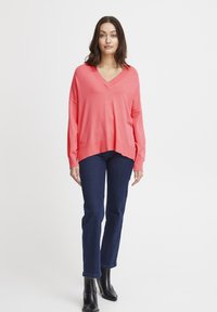 Maglione oversized a V in corallo con spalle scese, abbinato a jeans dritti blu scuro e stivaletti neri. Design minimalista, texture liscia.