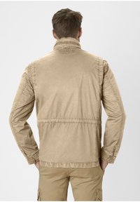 Beige cotton jacket with a stand-up collar, elastic waist, and long sleeves; features a textured finish and discreet branding on the collar.