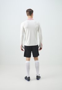 Vit långärmad träningsöverdel med ribbad textur och rosa krage, matchad med svarta shorts och vita strumpor, sedd från baksidan.