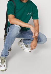 Zelené bavlněné tričko s bílým nápisem "Young Boy", doplněné o světle modré opotřebované džíny a bílé tenisky s olivovými akcenty.