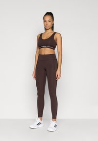 Brun sportstop med hvidt logo på banden, parret med matchende højttaljede leggings. Model iført hvide træningssko.