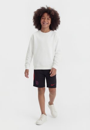 Smilende barn, der går, iført en hvid sweatshirt, sorte Nike sportsshorts med røde accenter og hvide sneakers mod en ensfarvet hvid baggrund.
