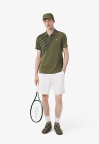 Olijfgroene polo shirt met een ruitpatroon, witte shorts en olijfkleurige sneakers. Accessoire omvat een bijpassende pet en een tennisracket.