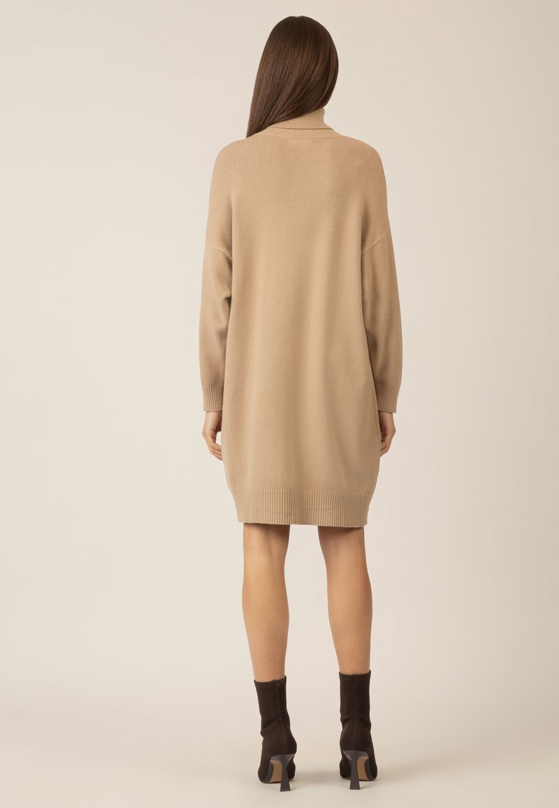 Frau mit langen braunen Haaren, die ein beige Knielanges Strickkleid und dunkelbraune Stiefel mit Absatz trägt, steht mit dem Rücken zur Kamera.