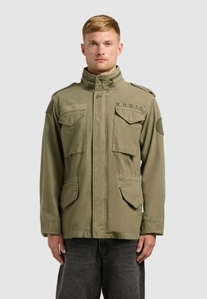 Mladý muž so svetlými krátkymi vlasmi, oblečený v olivovo zelenej vojenskej bunde s nápisom „K.H.U.J.O.“ a tmavosivých nohaviciach, stojaci nehybne.