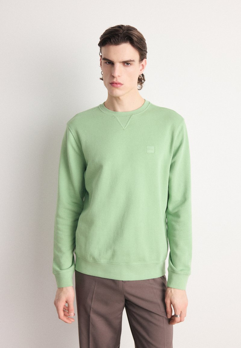Groene sweatshirt van zacht materiaal, ronde hals, ribgebreide manchetten en zoom, met een klein logo op de borst, gecombineerd met een paar bruine broek.
