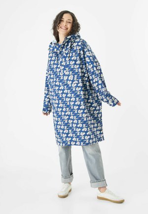 Blauwe waterdichte regenjas met witte cartoonpatronen, voorzien van een capuchon, lange mouwen en een losse pasvorm, gecombineerd met lichte spijkerbroeken en witte sneakers.