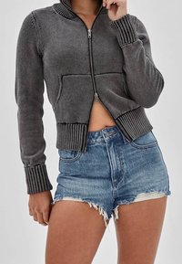 Szary sweter rozpinany na zamek z dzianiny, z prążkowanym kołnierzem i mankietami oraz przednią kieszenią. Do kompletu dżinsowe szorty z przetarciami.