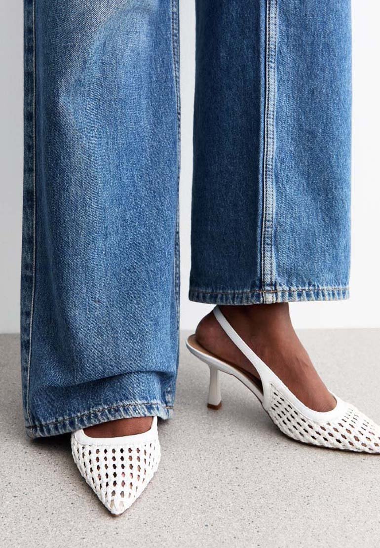 Vita spetsiga slingback klackar med ett vävt mönster, i kombination med ljusblå vida jeans. Platt, texturerad yta under.