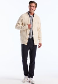 Chunky crèmekleurige cardigan met kabelbreimotief, houten knopen, gedragen over een blauw-wit gestreept shirt, gecombineerd met donkerblauwe broek en witte sneakers.