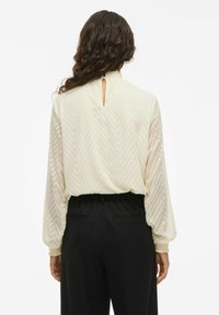 Light cream blouse with a chevron pattern, long sleeves, elastic cuffs, and a keyhole back detail, paired with black high-waisted pants.