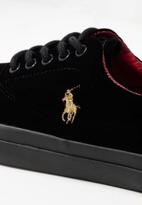 Musta samettinen lenkkari pyöreällä kärjellä, jossa on mustat nauhat, kultaiset brodeeratun hevosen logo ja musta kumipohja. Punainen kuviollinen vuoritus näkyy.