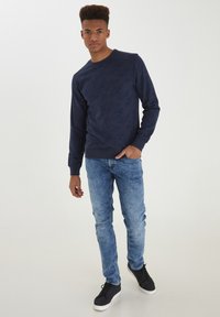 Felpa blu navy con motivo di foglie in rilievo, collo a giro, e polsini a costine. Abbinata a jeans denim azzurri e sneaker nere.