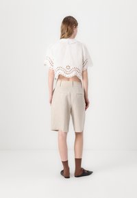 Biely crop top s očkovaným vzorom a vlnitým lemom a krátkymi rukávmi, kombinovaný so svetlo béžovými ľanovými šortkami. Hnedé ponožky a čierne topánky dotvárajú outfit.