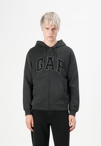 Jeune personne aux cheveux blonds courts portant un sweat à capuche zippé gris foncé de GAP et un pantalon noir, se tenant devant un fond blanc.