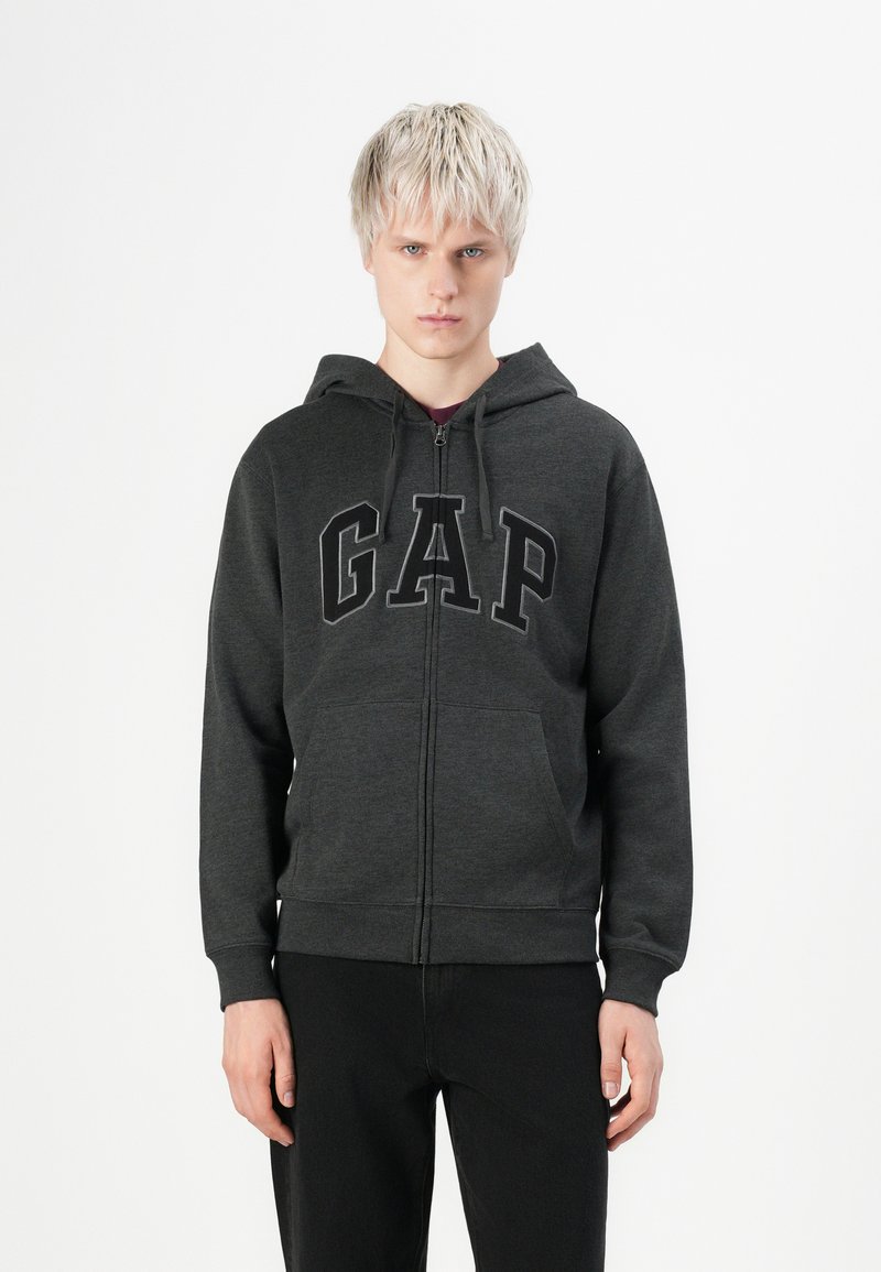 Jeune personne aux cheveux blonds courts portant un sweat à capuche zippé gris foncé de GAP et un pantalon noir, se tenant devant un fond blanc.