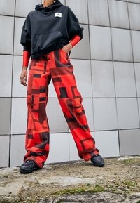 Felpa nera oversize con tasca frontale e logo, abbinata a pantaloni rossi con un motivo geometrico nero. Sneakers nere.