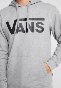 Pelēks kapucis izgatavots no mīksta auduma, ar lielu melnu "VANS" logo priekšpusē, kangaroopa kabatu un aukliņu kapuci.