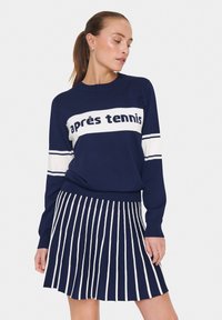 Marinblå tröja med vit "après tennis" text, i kombination med en veckad kjol med vertikala vita ränder. Enkel, atletisk design.