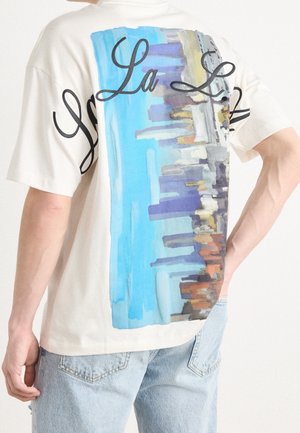 Person, die ein weißes T-Shirt mit einer blauen Stadtsilhouette und dem kursiven Text "La L." auf dem Rücken trägt, kombiniert mit hellblauen Jeans.