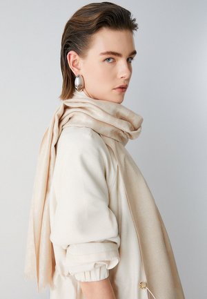Sciarpa di seta beige drappeggiata su un indumento di colore chiaro e dalla trama evidente. Grandi orecchini metallici in evidenza. Superficie liscia e lucida con motivi sottili.