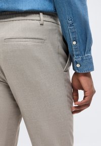 Άνδρας με μπεζ υφασμάτινο παντελόνι και μπλε τζιν πουκάμισο με κουμπωμένη μανσέτα, το χέρι χαλαρό στο πλάι, μπροστά σε ανοιχτό φόντο.