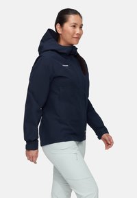 Giacca impermeabile blu navy con cappuccio, dotata di due tasche frontali e polsini elastici. Indossata sopra pantaloni di colore chiaro, con una texture liscia.