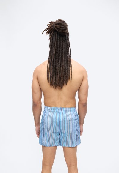 Άνδρας με μακριά κοτσίδα κατάλληλα δεμένη, χωρίς πουκάμισο, φοράει μπλε και λευκές ριγωτές βερμούδες, στέκεται με την πλάτη στον θεατή πάνω σε απλό λευκό φόντο.