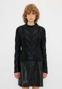 Schwarzer Strickpullover mit erhöhtem Kabelmuster, geripptem Saum und Bündchen. Getragen über einem glänzenden schwarzen Lederrock, der bis zur Mitte des Oberschenkels reicht.
