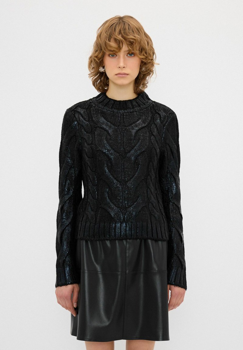 Schwarzer Strickpullover mit erhöhtem Kabelmuster, geripptem Saum und Bündchen. Getragen über einem glänzenden schwarzen Lederrock, der bis zur Mitte des Oberschenkels reicht.