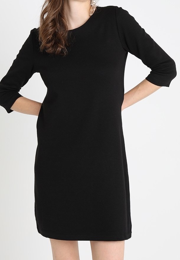 Femme portant une robe noire simple longueur genou avec des manches trois-quarts, posant les mains sur les hanches devant un fond uni.