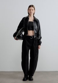 Μαύρο δερμάτινο cropped μπουφάν με ψηλό γιακά πάνω από μαύρο cropped τοπ με κουμπιά, συνδυασμένο με φαρδιά μαύρη παντελόνα και χοντρά παπούτσια.