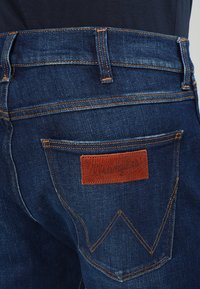Gros plan de la poche arrière d'un jean denim bleu foncé avec des surpiqûres orange et un écusson logo Wrangler en cuir marron.