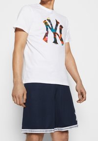 T-shirt en coton blanc avec un logo coloré de New York, associée à un short bleu marine avec des rayures blanches et un logo distinctif sur le côté.