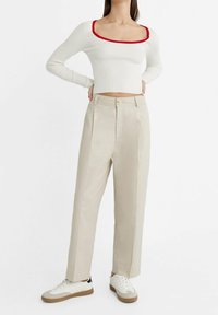 Žena v bílém dlouhém rukávu krátkém topu s červenými lemy, béžových kalhotách s vysokým pasem a bílých teniskách s hnědými podrážkami.